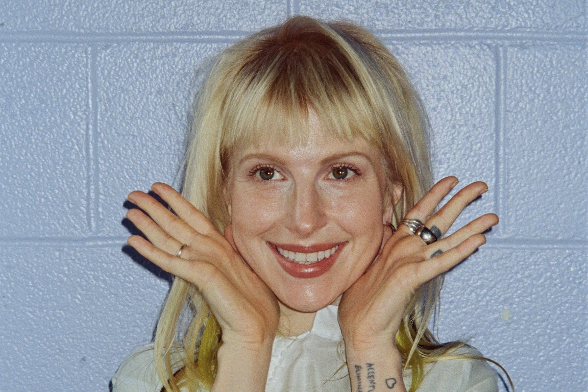 Singer Hayley Williams smiling with blond hair and bangs, framing her face with hands open against a textured blue cinder block wall.
